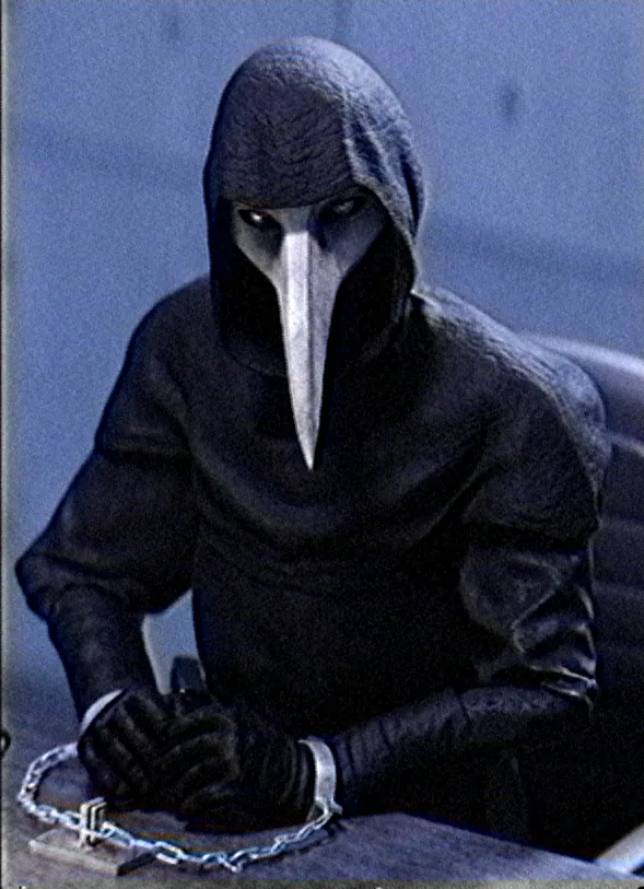 A figure in a dark hooded robe and a long-beaked mask sits with chained hands folded on a table.